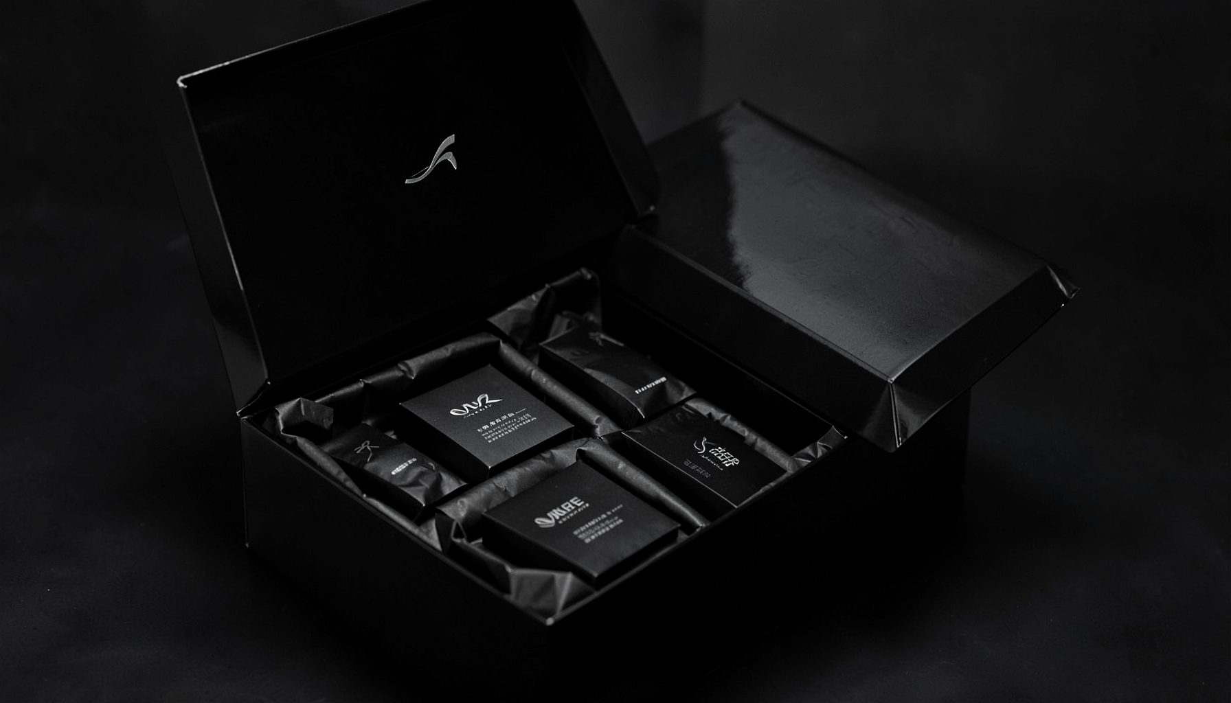 A glossy black beauty box with a sleek logo, opened to reveal neatly arranged products in black tissue, sophisticated and dramatic mood.
